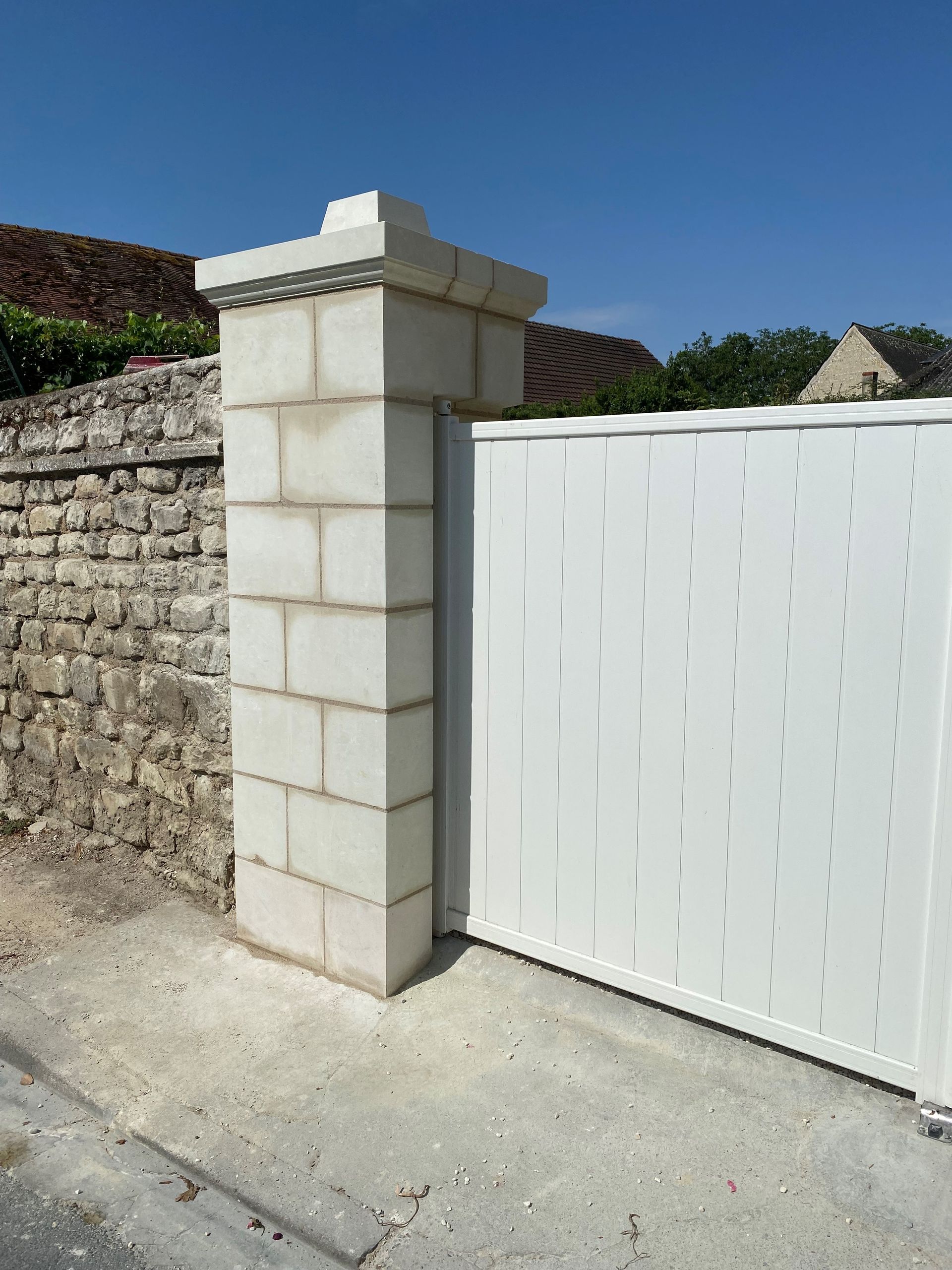 Portail blanc à côté d'un pilier en parpaings surmonté d'un chapiteau carré.
