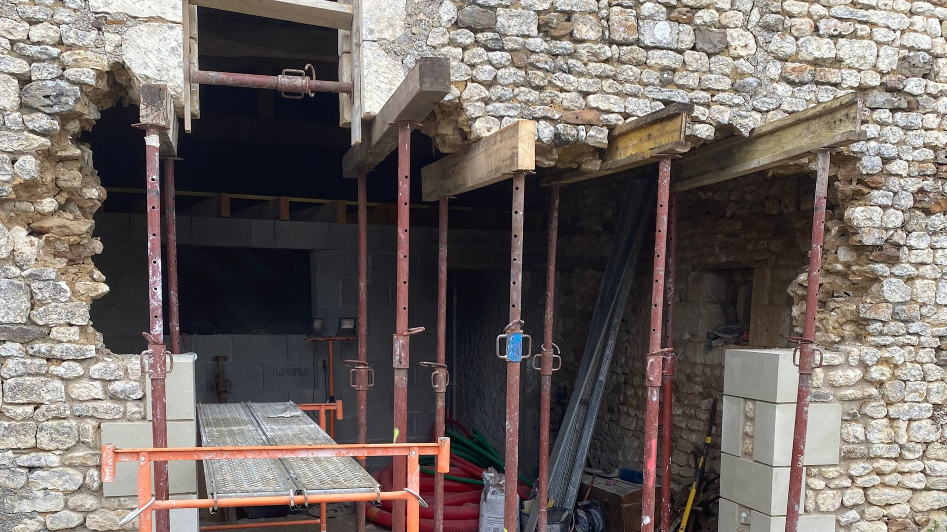 Extérieur d'un bâtiment en construction avec un mur en pierre apparente, soutenu par des poutres métalliques et en bois.