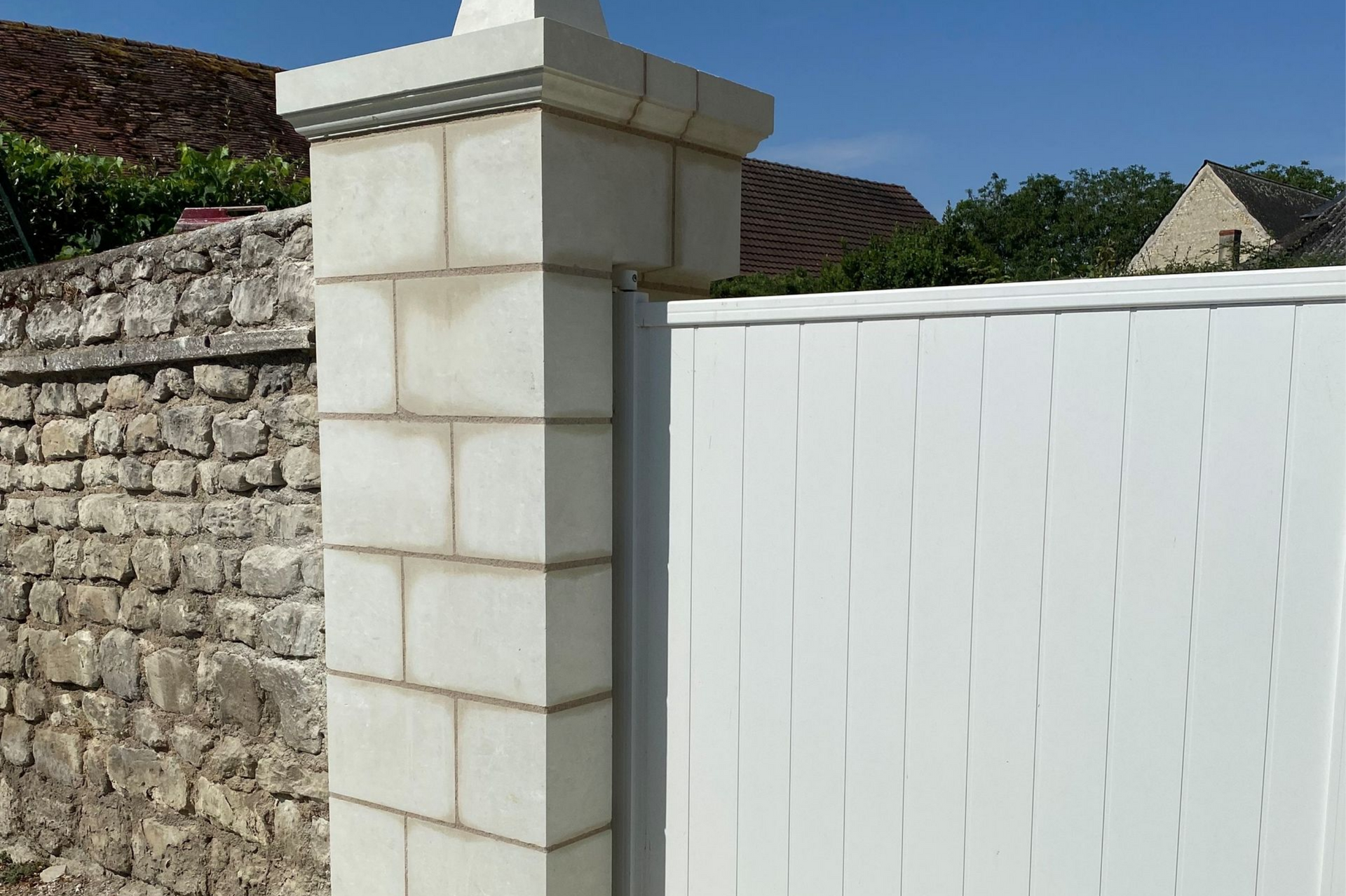 Colonne de pierre à chapiteau décoratif, à côté d'une clôture blanche et d'un mur de pierre.