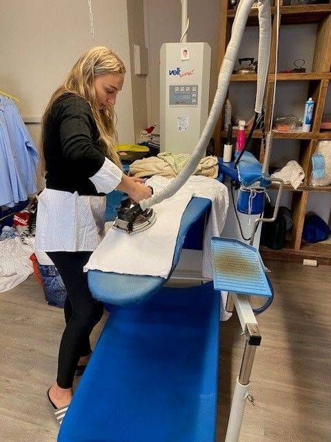 Eine Frau bügelt in einem vollgestellten Raum mit einem Dampfbügeleisen Kleidung an einer Wäschereistation.