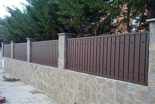 Un muro de piedra con una valla metálica y árboles al fondo.