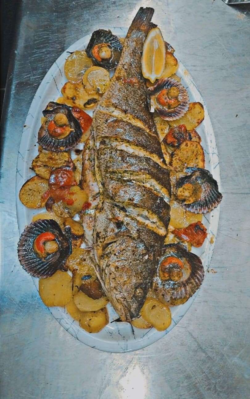 Plato de pescado entero a la parrilla adornado con rodajas de limón y verduras en una bandeja blanca.