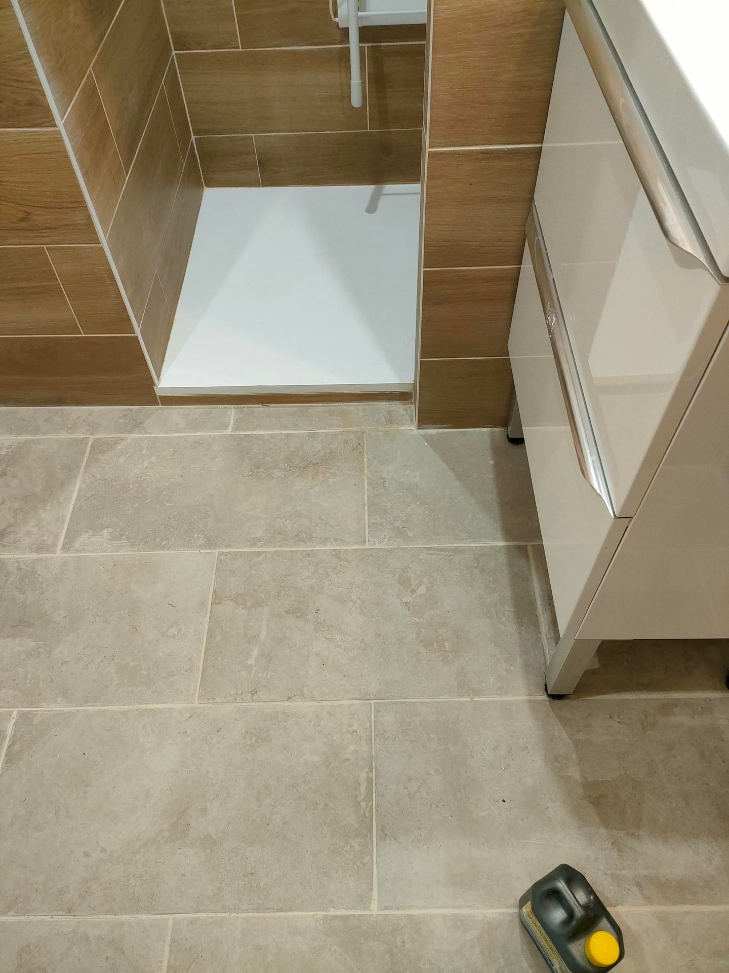 Salle de bains avec sol carrelé beige, murs carrelés imitation bois, receveur de douche blanc et meuble blanc.