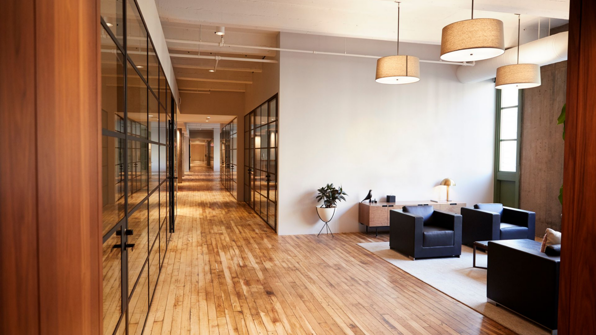Un intérieur de bureau moderne avec parquet, couloirs vitrés et un coin salon avec fauteuils noirs.