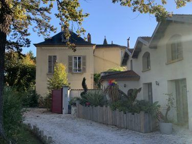 Bâtiments aux murs clairs et aux toits sombres, avec une clôture en bois et un chemin de gravier devant.