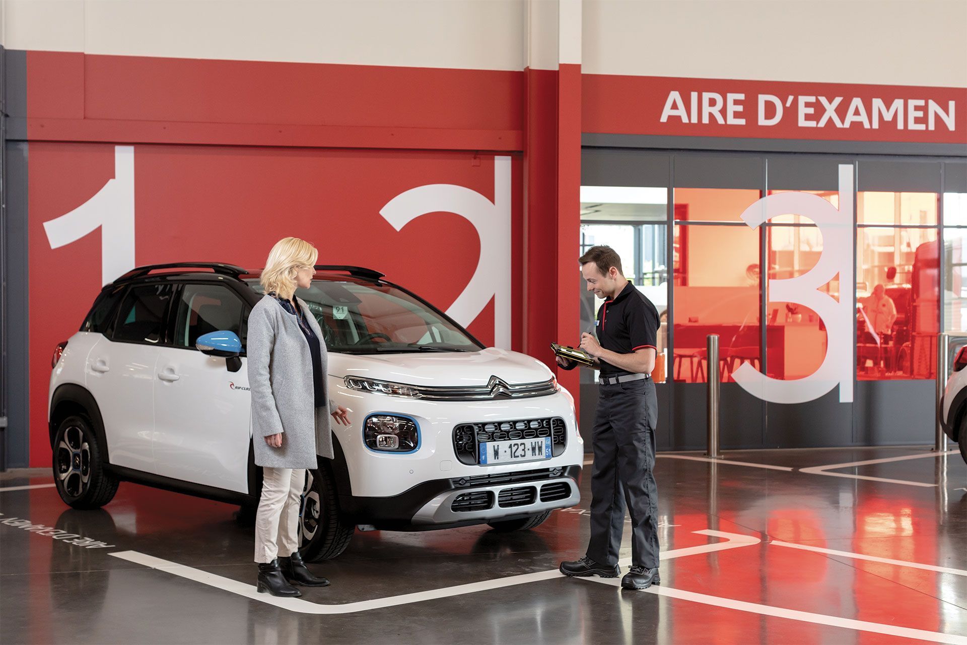 Un homme et une femme près d'une voiture blanche devant le panneau Aire d'examen. L'homme tient un appareil.
