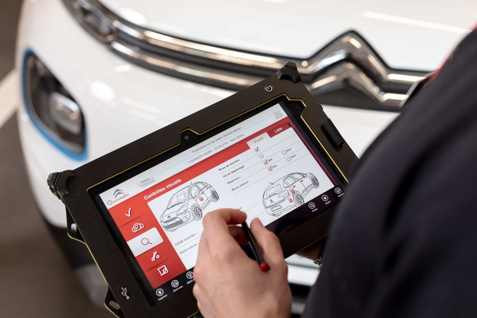 Une personne utilise une tablette pour diagnostiquer une voiture blanche. La tablette affiche un schéma du véhicule.