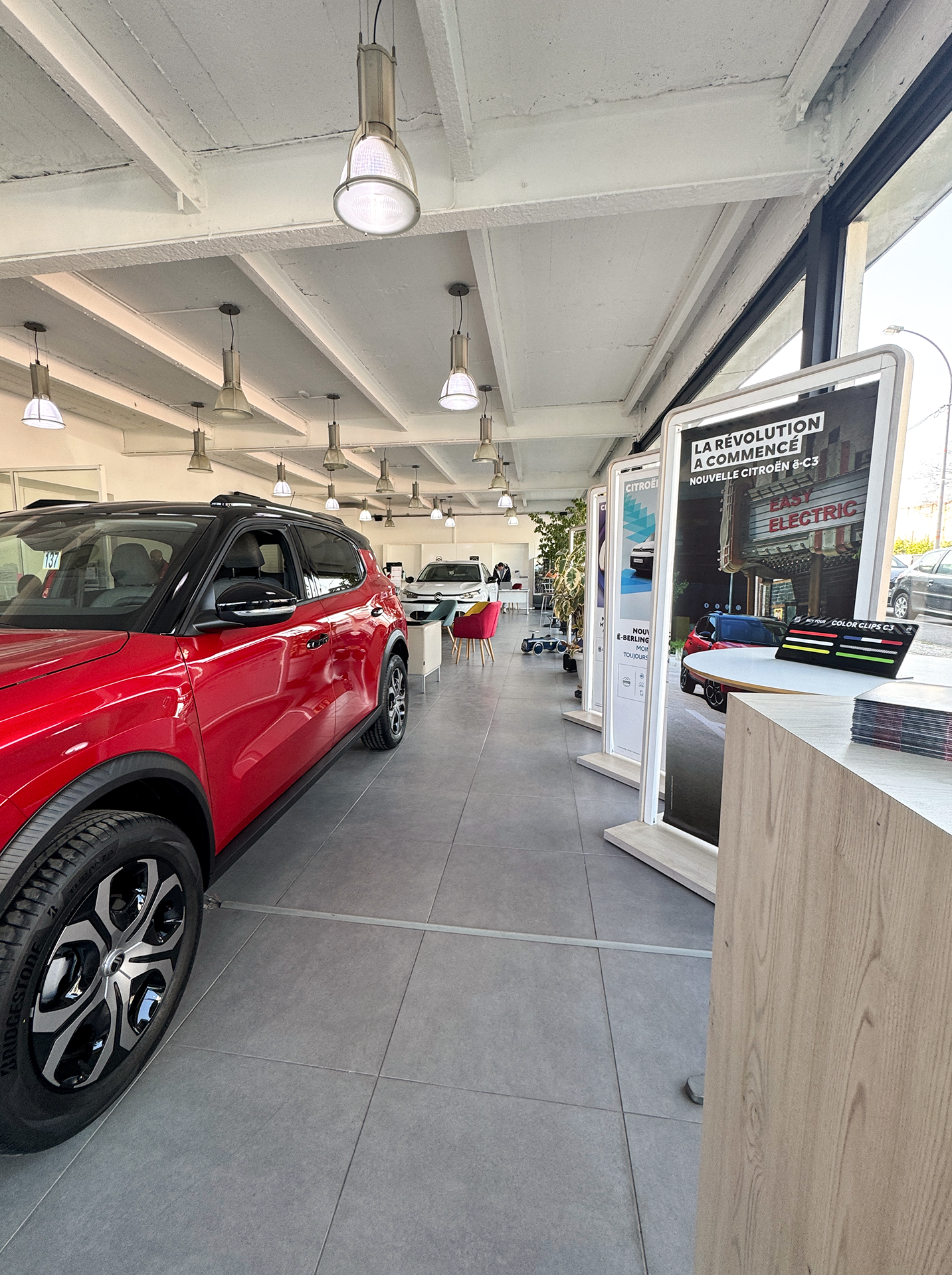 Voiture rouge garée à l'intérieur d'une concession automobile, avec des panneaux d'information et un éclairage au plafond.