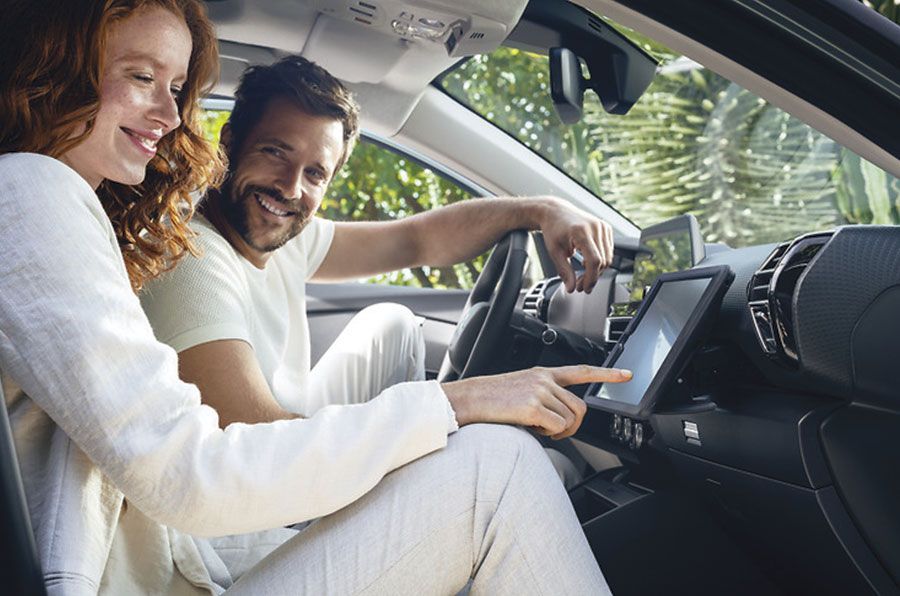 Un couple souriant dans une voiture ; l’homme pointe du doigt un écran tactile, la femme le regarde. Lumière du soleil, vue intérieure.