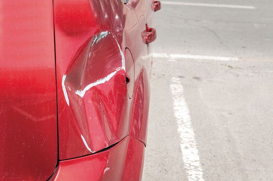 Panneau latéral arrière d'une voiture rouge présentant un gros enfoncement et des rayures sur la peinture, garée près d'une ligne blanche de stationnement.