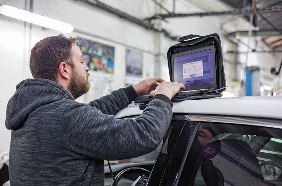 Un mécanicien utilise un ordinateur portable sur le toit d'une voiture dans un garage, en train de taper et d'inspecter l'écran.