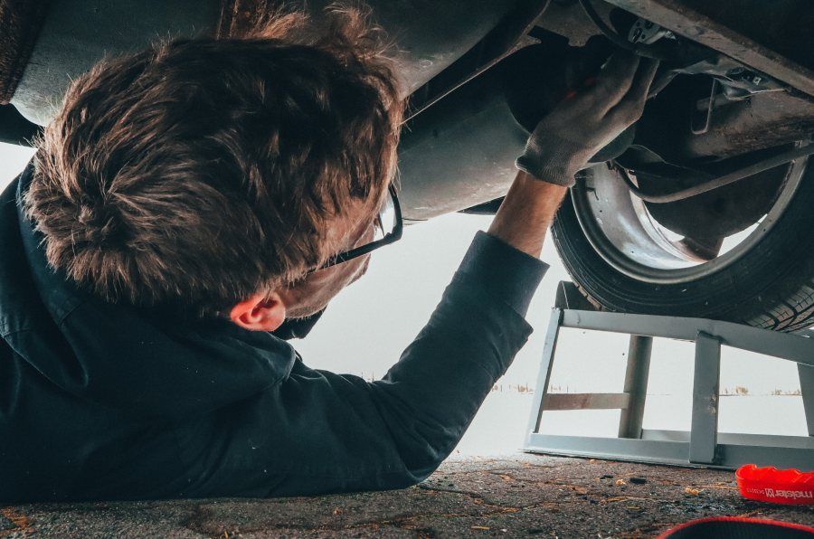 Une personne travaille sous la voiture, portant des lunettes et des gants. La voiture est surélevée sur des parpaings.
