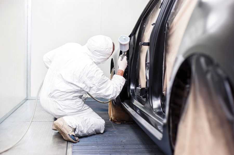 Une personne en combinaison de protection peint une portière de voiture à l'intérieur d'une cabine de peinture.