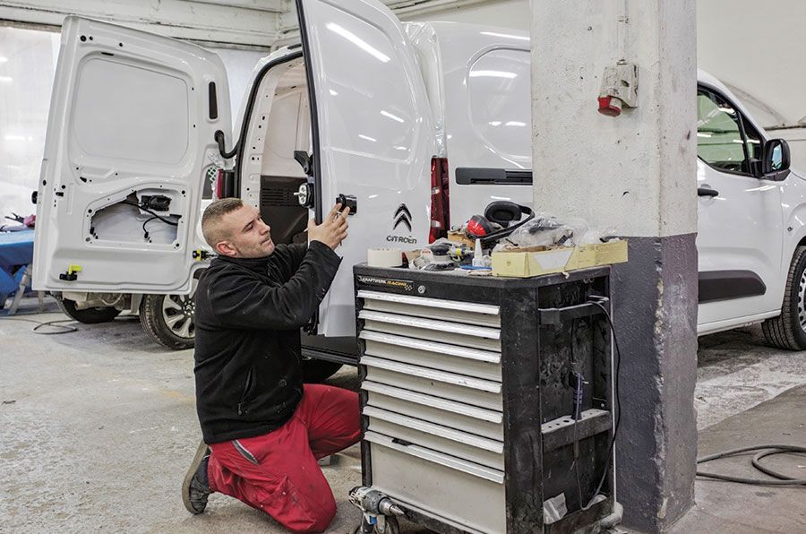 Un mécanicien travaille à l'arrière d'une camionnette blanche dans un garage, utilisant un outil en étant agenouillé.