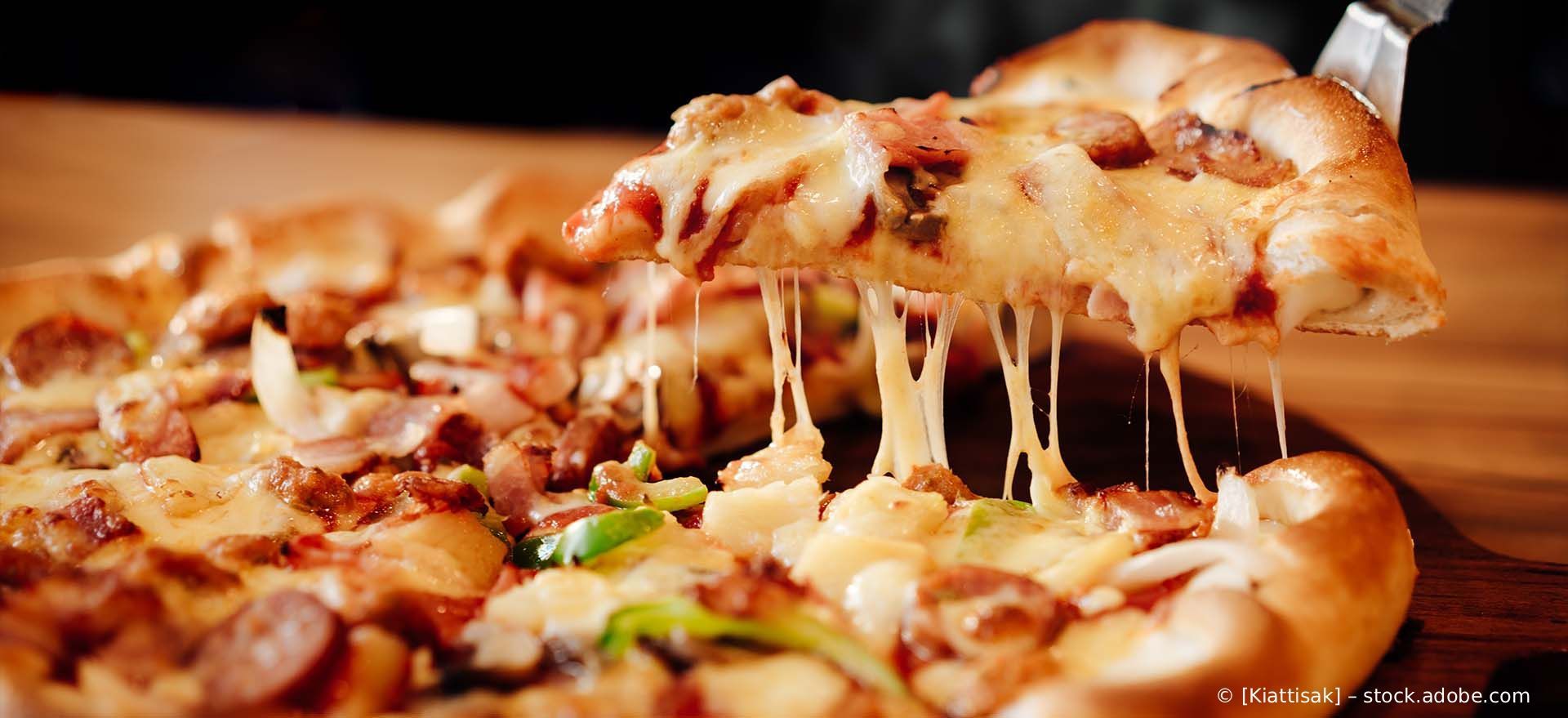 Ein Stück reichhaltig belegte Pizza wird mit einem Pfannenwender angehoben, wobei Fäden aus geschmolzenem Käse über einen Holztisch verlaufen.