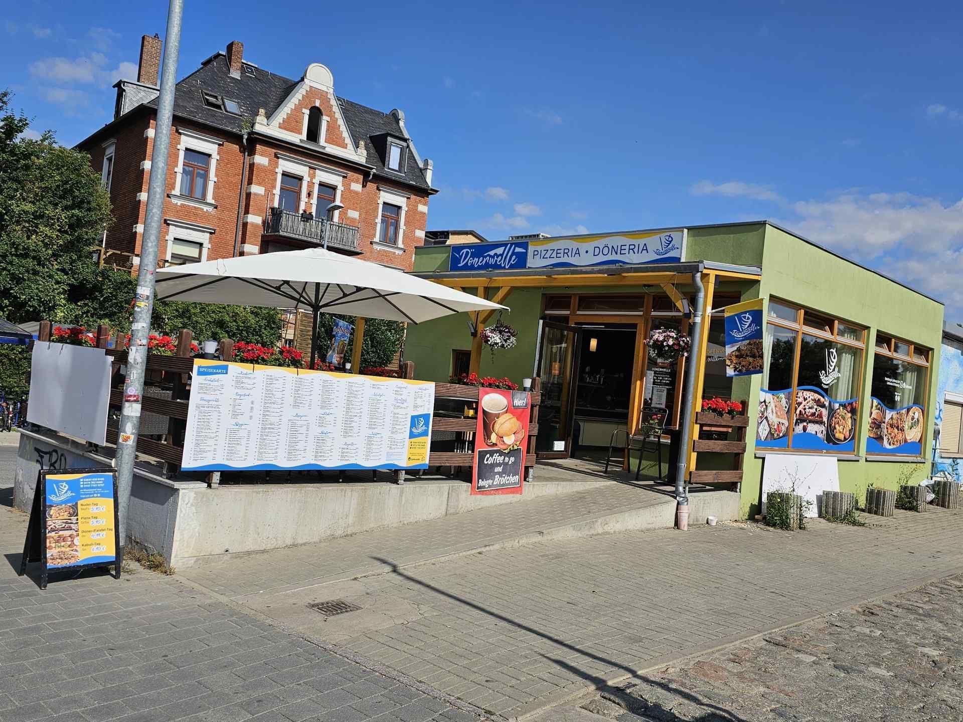 Ein hellgrünes Restaurantgebäude mit Außensitzplätzen, einem großen Speisekartenschild und einem traditionellen Backsteinhaus im Hinterhof.