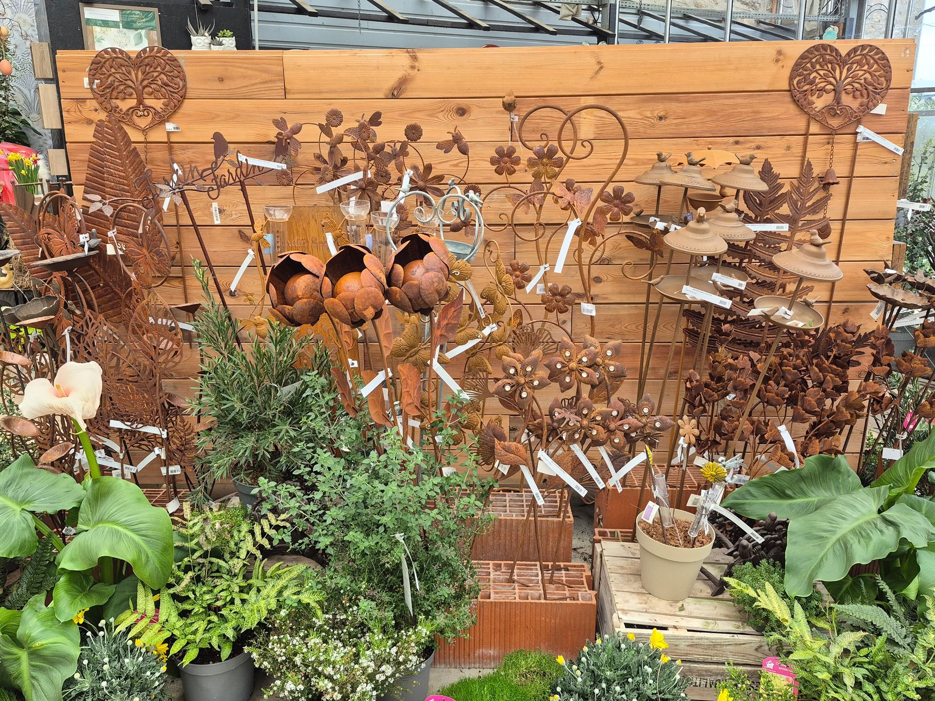 Dans une pépinière, divers tuteurs de jardin en métal rouillé sont disposés contre un mur en bois, entourés de plantes vertes en pot.