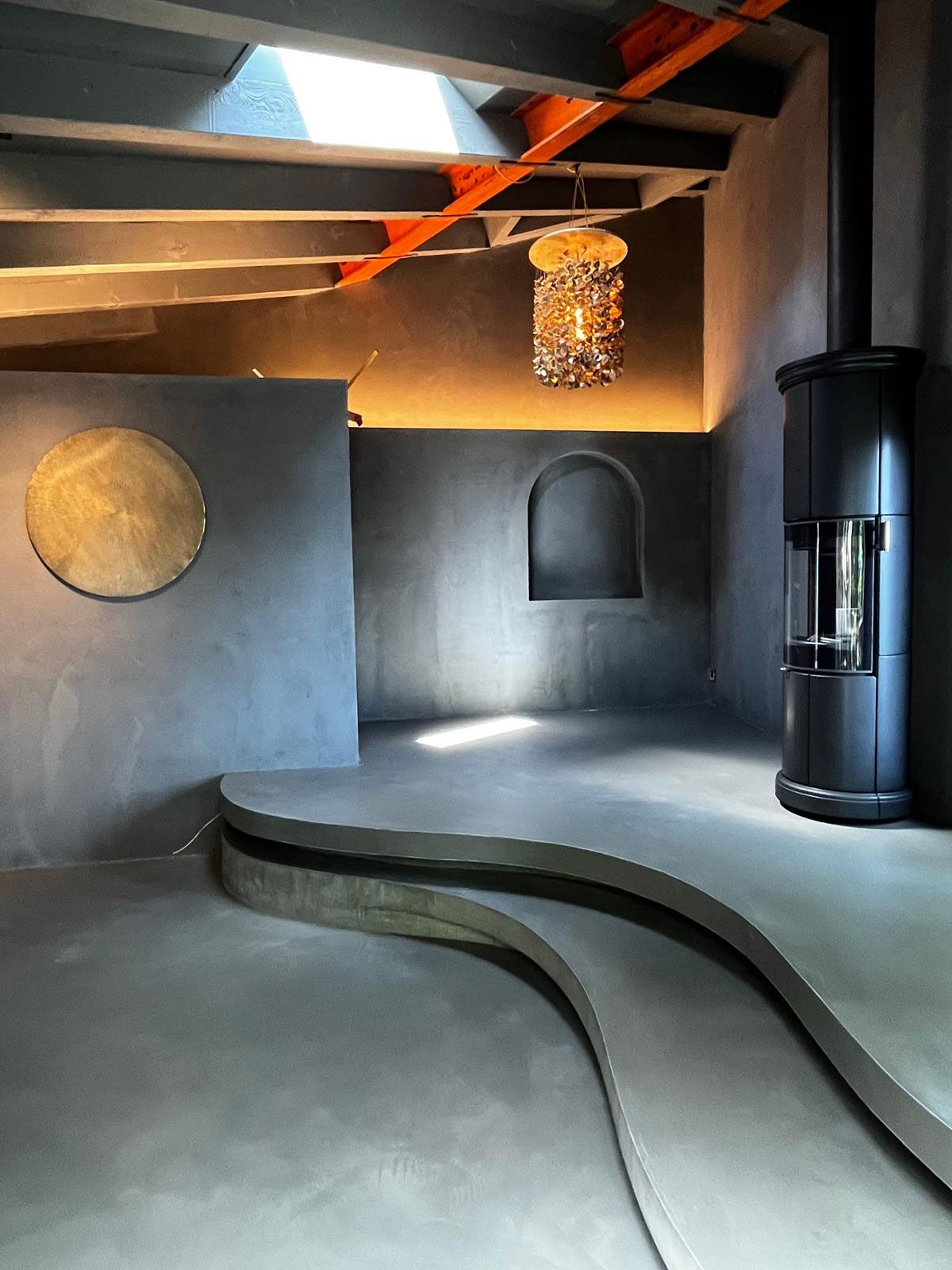 Ein minimalistischer Raum im Beton-Stil mit einer geschwungenen Plattform, einem schwarzen Herd, einem goldenen runden Wandelement und einer Pendelleuchte.