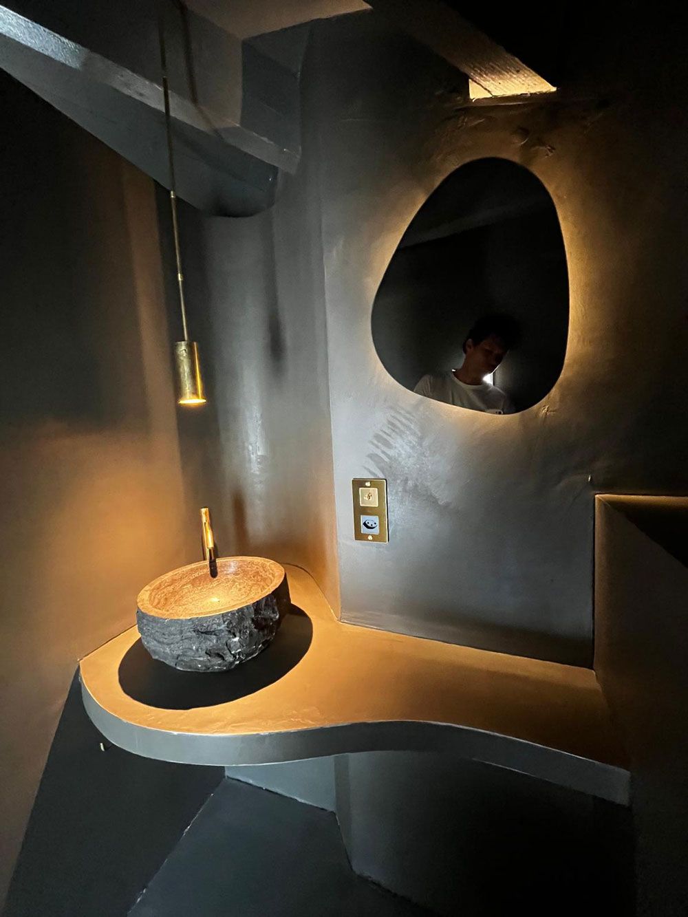 Ein moderner Badezimmerwaschtisch mit geschwungener Betonplatte, einem Aufsatzwaschbecken aus Stein, einem goldenen Wasserhahn und einem hinterleuchteten, unregelmäßigen Spiegel.