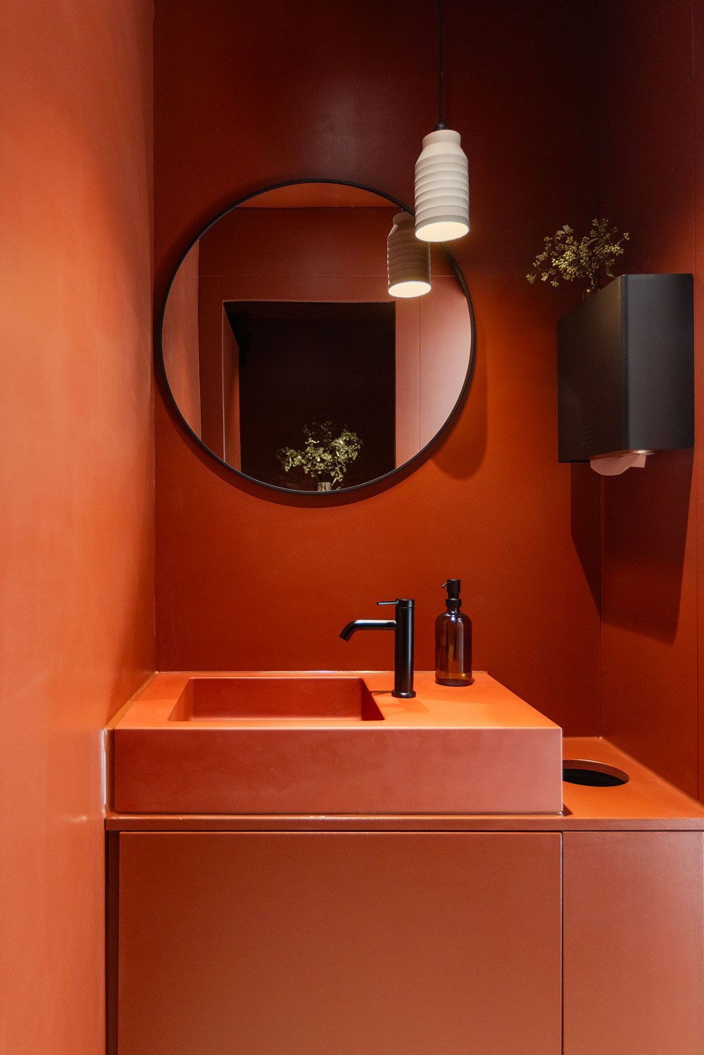 Ein minimalistisches Badezimmer mit terrakottafarbenen Wänden, einem passenden Waschbecken, einem runden Spiegel und einer weißen Hängeleuchte.