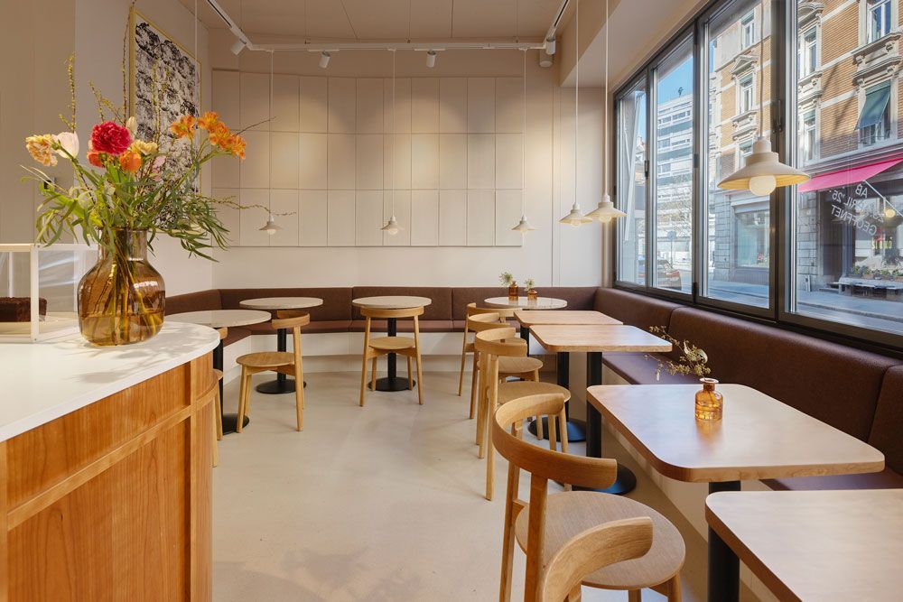 Ein helles, minimalistisches Café-Interieur mit Holztischen, braunen Sitzbänken, hellen Holzstühlen und einer Blumenvase.