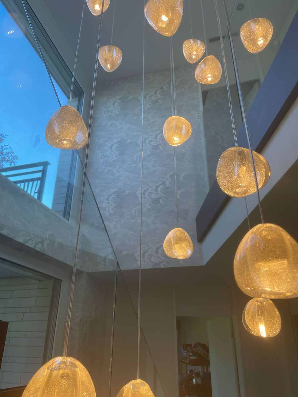 In einem modernen, mehrgeschossigen Innenraum hängt eine Gruppe leuchtender, strukturierter Glaspendelleuchten in unterschiedlichen Höhen.