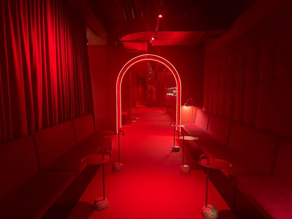 Ein in rotes Licht getauchter Raum mit einem neonbeleuchteten Torbogen, einem rot ausgelegten Weg und passenden Samtsitzen auf beiden Seiten.
