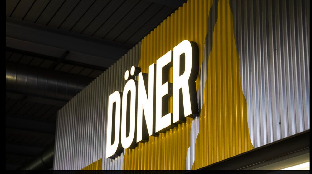 Das Wort „DÖNER“ in großen, weißen Leuchtbuchstaben auf einer gelb-grauen Wellblechwand.