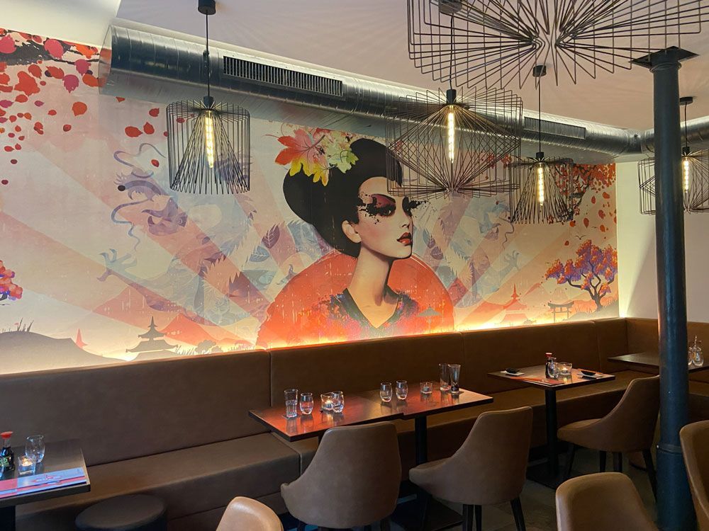 Das Interieur eines Restaurants zeigt ein Wandgemälde einer Figur mit floralem Haarschmuck, braune Sitznischen und Metallbeleuchtung.