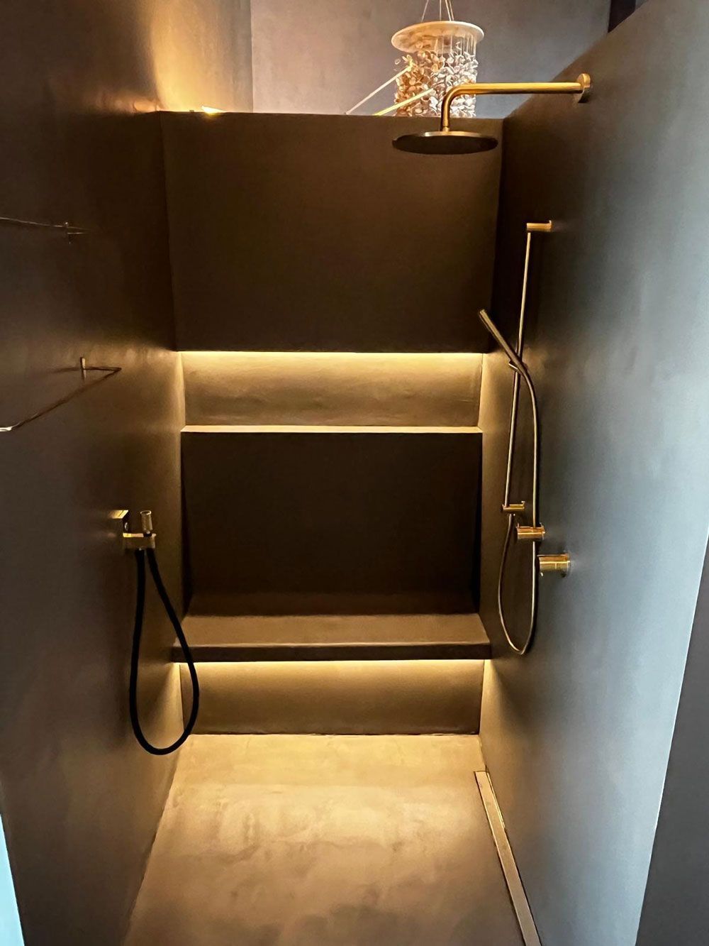 Eine moderne, begehbare Dusche mit dunkelmatten Wänden, goldenen Armaturen, einer eingebauten Sitzbank und integrierter LED-Akzentbeleuchtung.