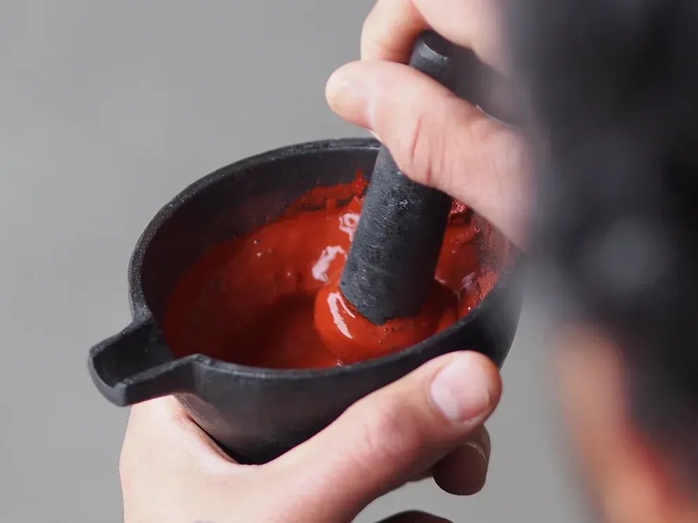 Eine Person hält einen Mörser und Stößel über eine Schüssel mit roter Soße.
