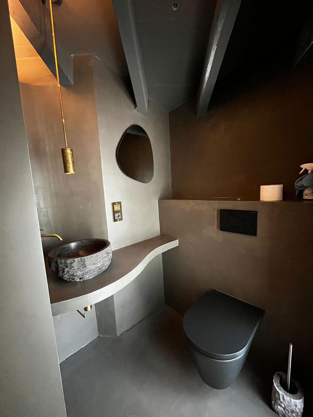 Minimalistisches Badezimmer mit Steinwaschbecken, organisch geformtem Spiegel, dunkler, wandhängender Toilette und gedeckten Erdtönen.