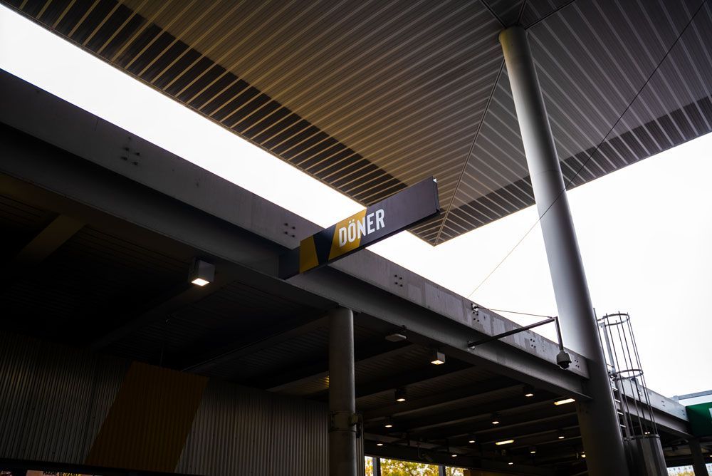 Ein Schild, das an der Betondecke eines Bahnhofs oder Terminals hängt und die Aufschrift „DONER“ in weißer Schrift zeigt.
