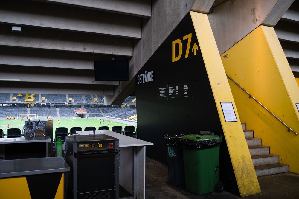Ein Stadionumlauf mit einer schwarzen Wand mit der Aufschrift „D7“ und einer Treppe, der auf ein Fußballfeld mit Sitzplätzen und einem Imbissbereich blickt.