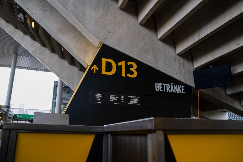Unter einer Betontreppe im Stadion befindet sich ein schwarzes Wegweiserschild mit gelber Aufschrift „D13“ und „GETRÄNKE“.