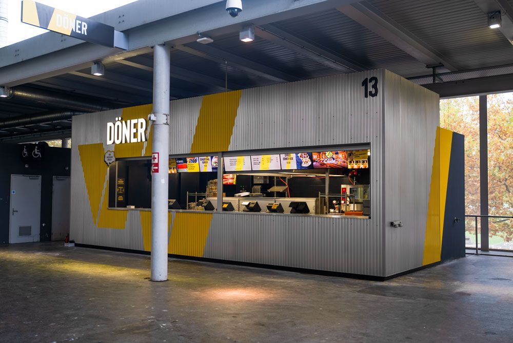 Ein moderner, grau-gelber Dönerstand mit der Nummer 13, der sich in einem Bahnhof befindet und über ein großes Ausgabefenster verfügt.