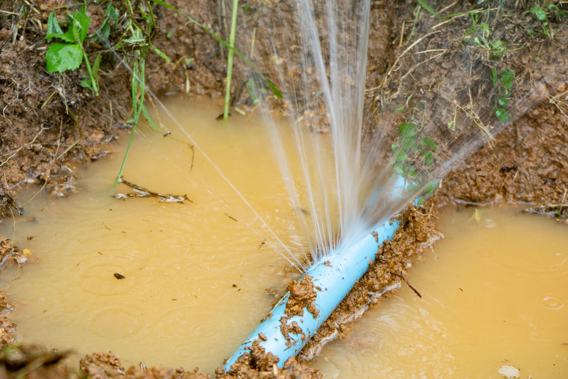 Compteur d'eau à couvercle bleu ouvert, laissant apparaître les cadrans et la vanne. Installé dans une zone de terre brune jonchée de débris.