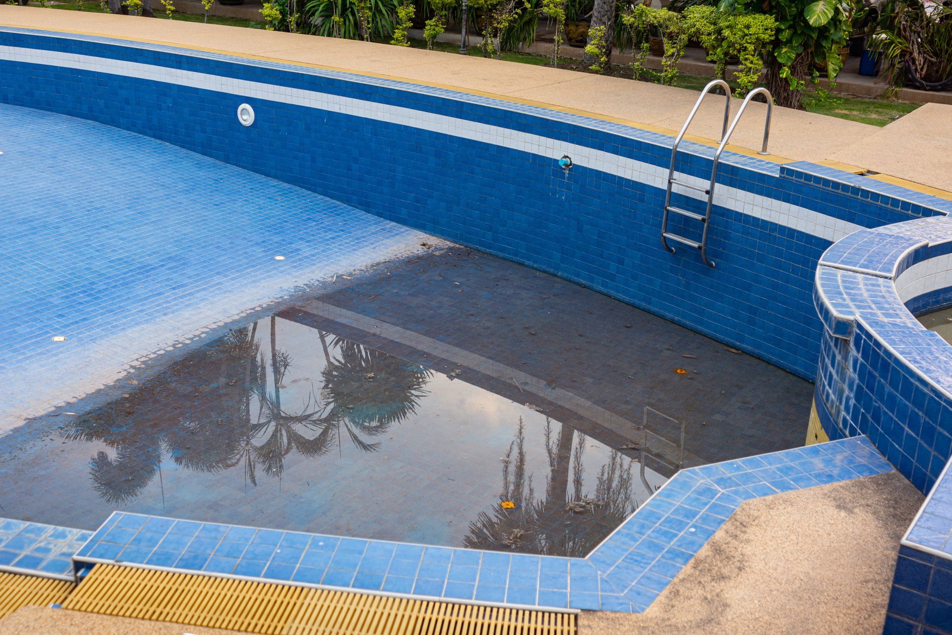 Piscine partiellement remplie, carrelée de bleu ; échelle et terrasse environnante.