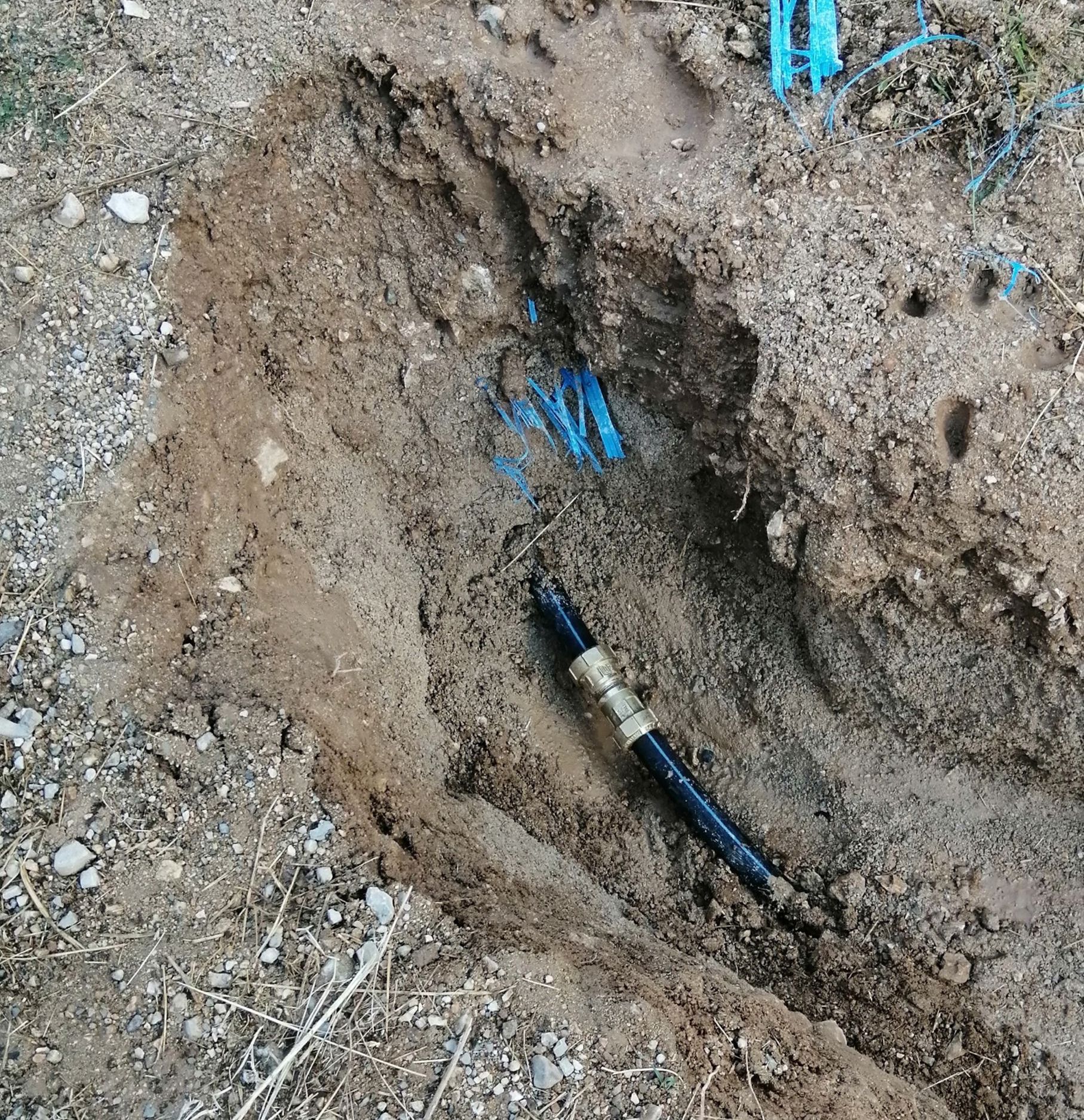Une tranchée creusée dans la terre révèle un tuyau noir avec des raccords en laiton et des conduites d'irrigation bleues.