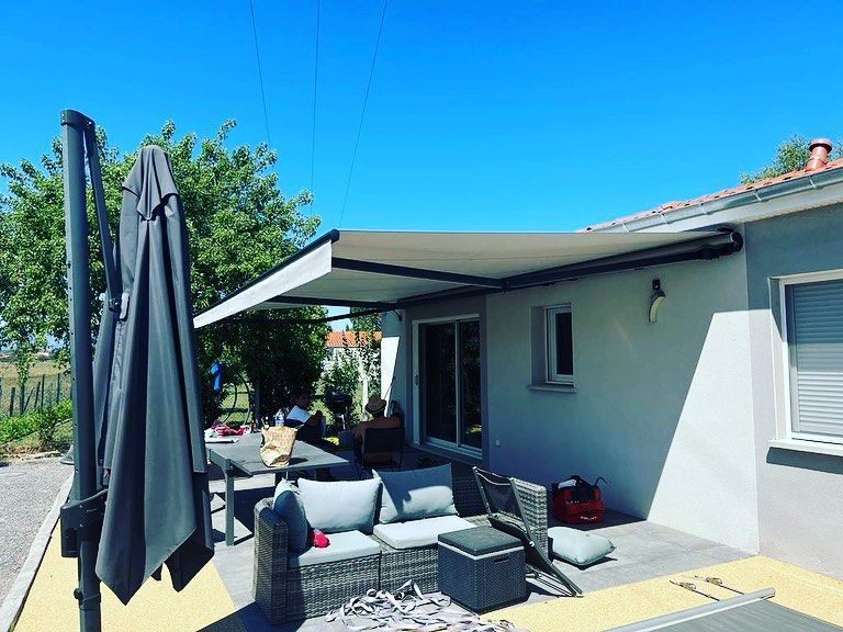 Terrasse meublée de table, chaises et sièges protégée du soleil par un grand store à banne.