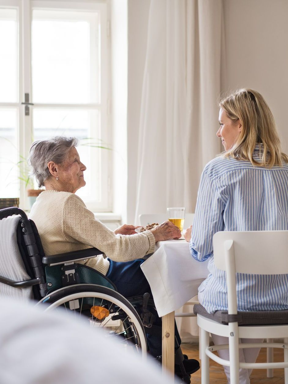 Une personne agée en fauteuil roulant discute avec une professionnelle assise à une table.