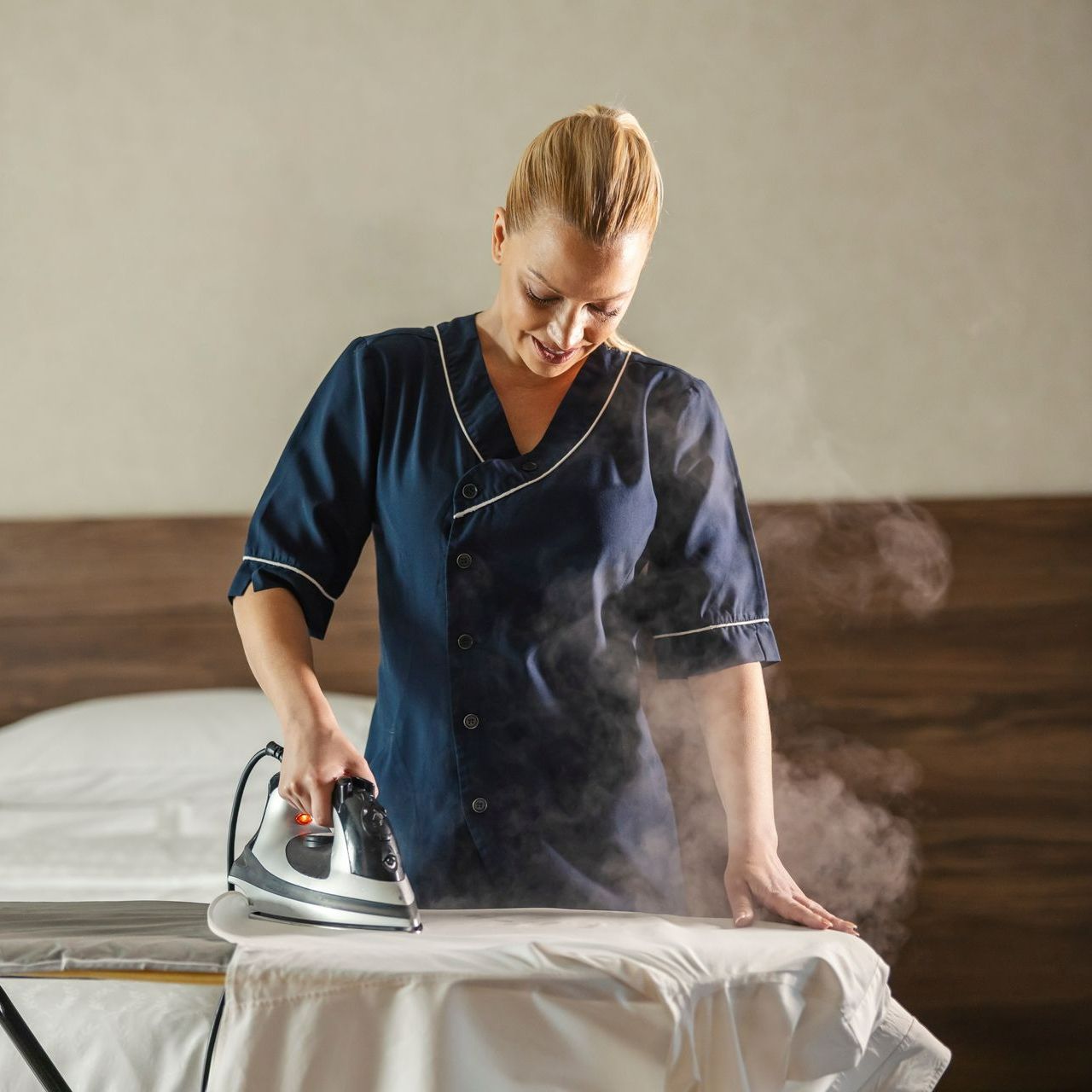 Une professionnelle repasse des draps devant un lit en arrière-plan.