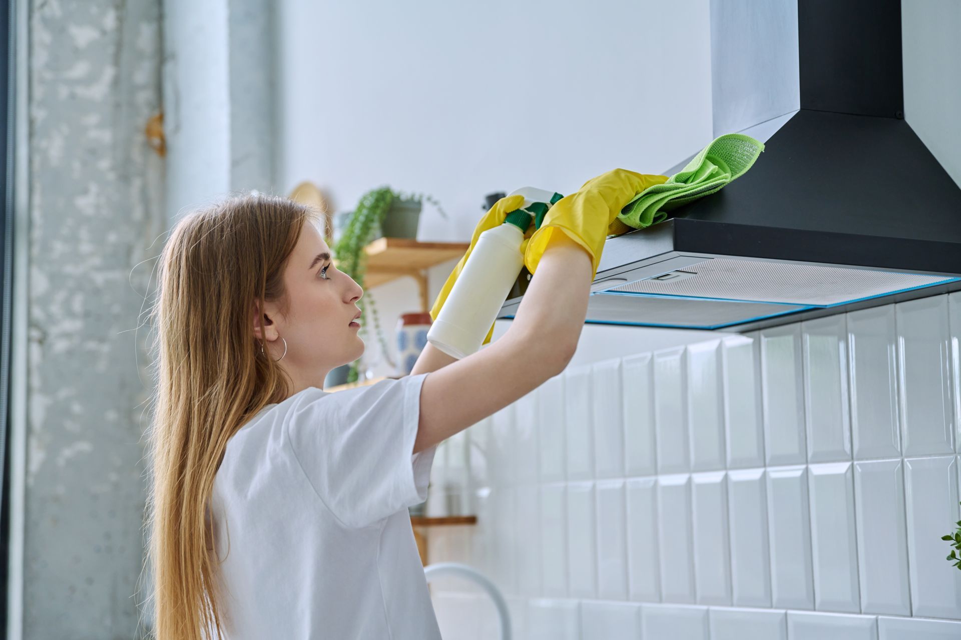 Une femme portant des gants nettoie la hotte de cuisine avec un vaporisateur et un chiffon.
