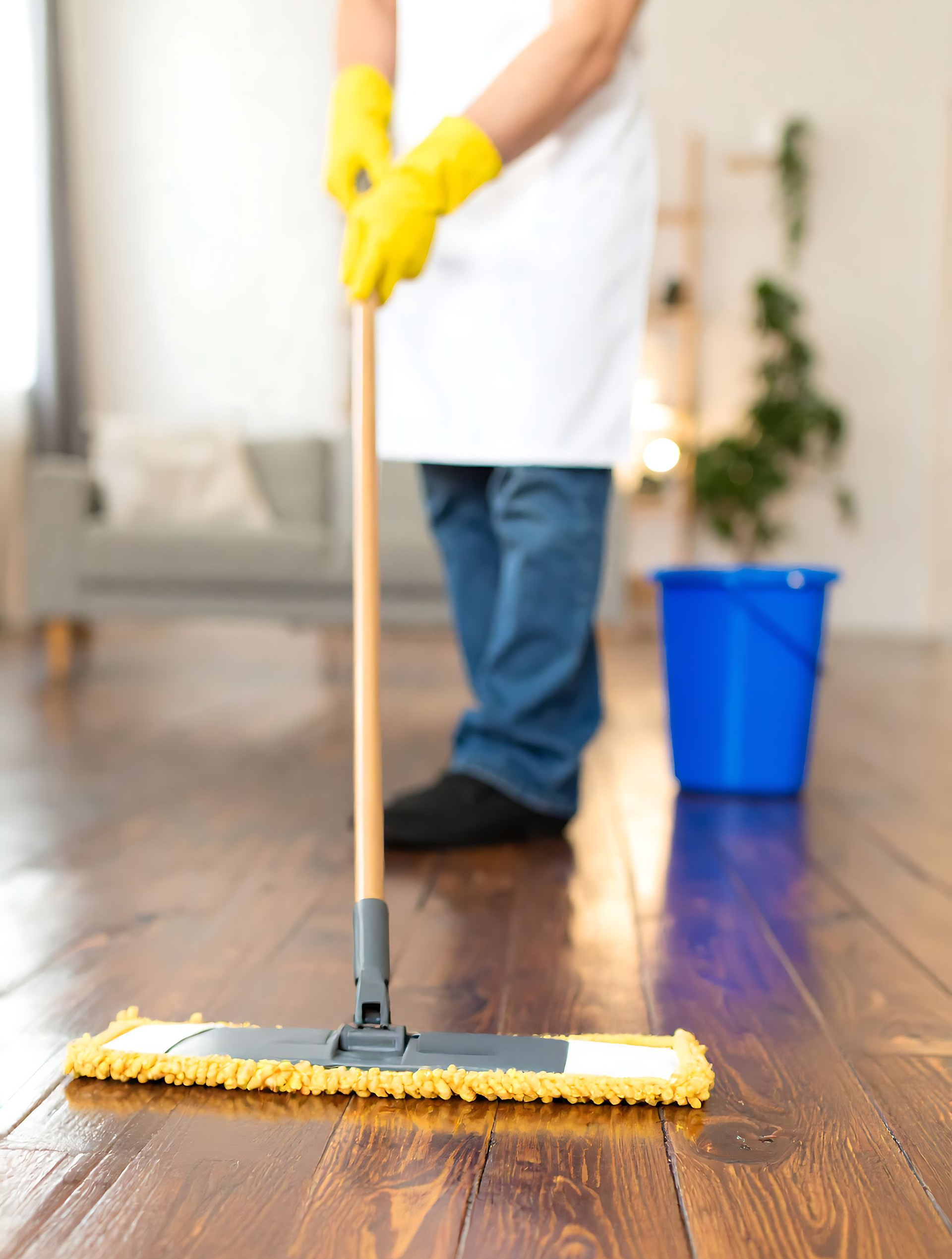 Personne nettoyant un plancher en bois avec une serpillière jaune, portant des gants jaunes et un seau bleu en arrière-plan.