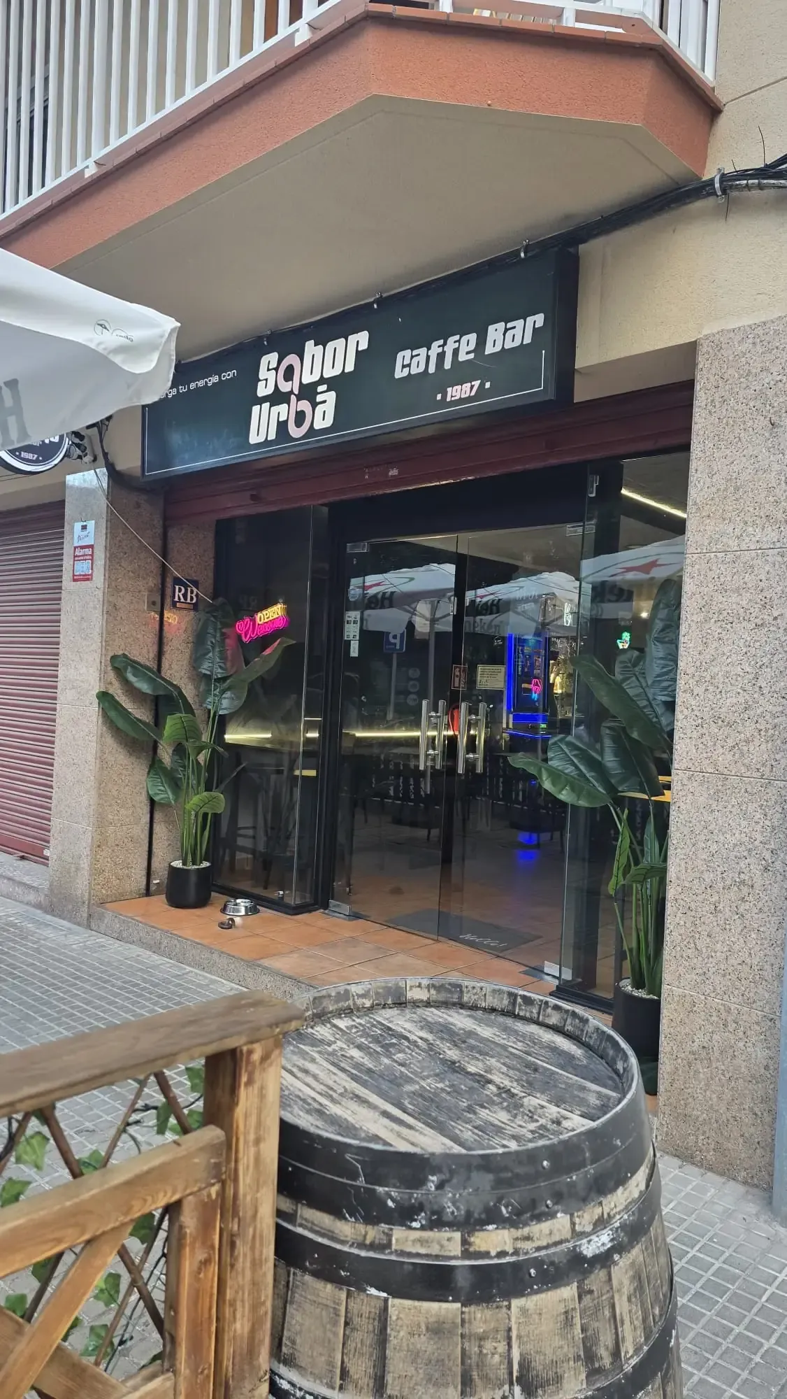 Interior de restaurante con carteles de comida de neón en una pared de madera y plantas; mesa y sillas en primer plano.