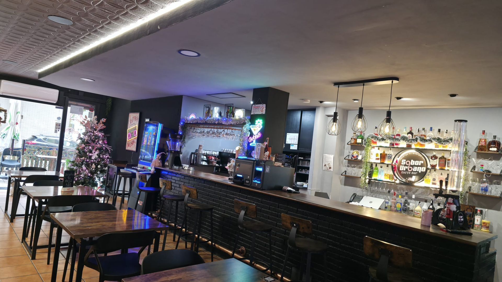 Entrada al bar cafetería “Sabor Urbà” con puertas de cristal, plantas en macetas y un interior oscuro.