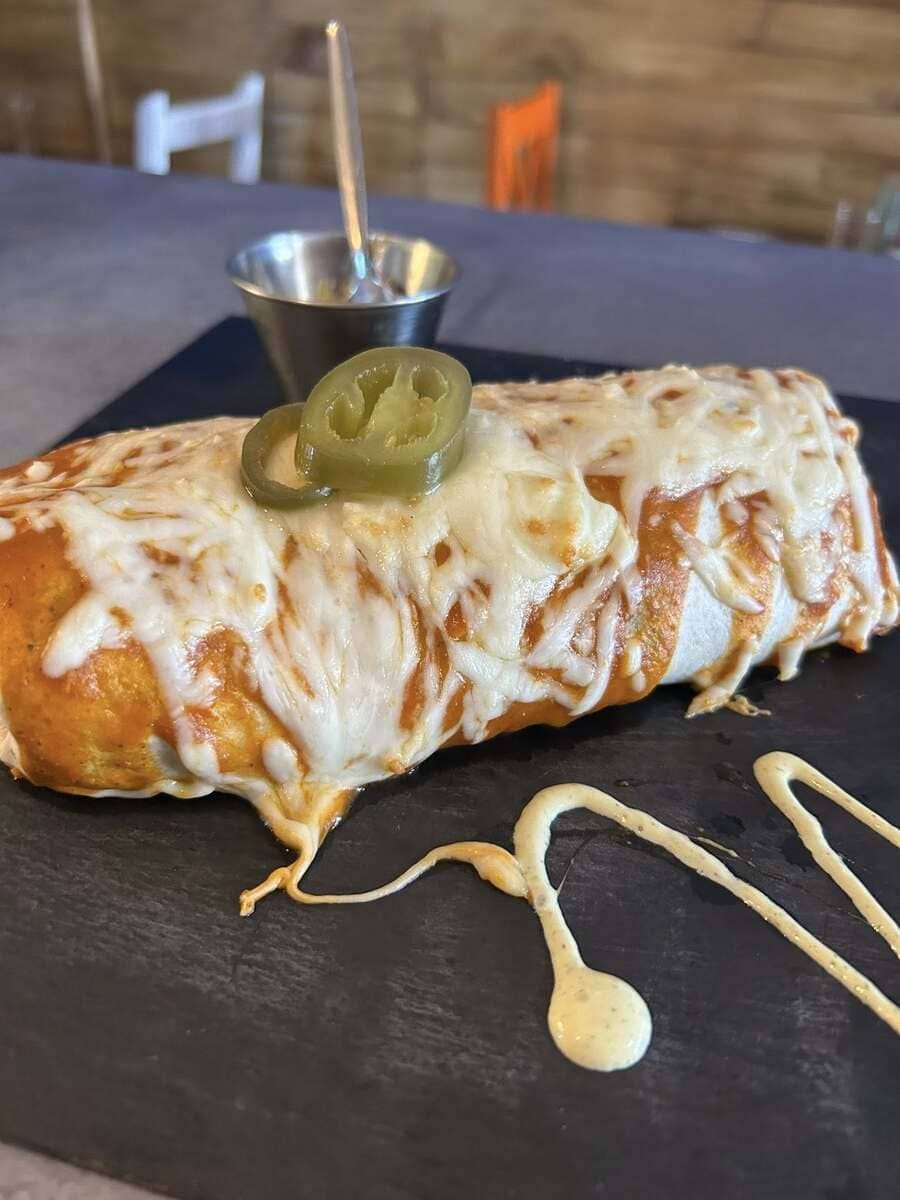 Burrito cubierto de queso derretido, cubierto con rodajas de jalapeño, rociado con salsa, servido en pizarra negra.