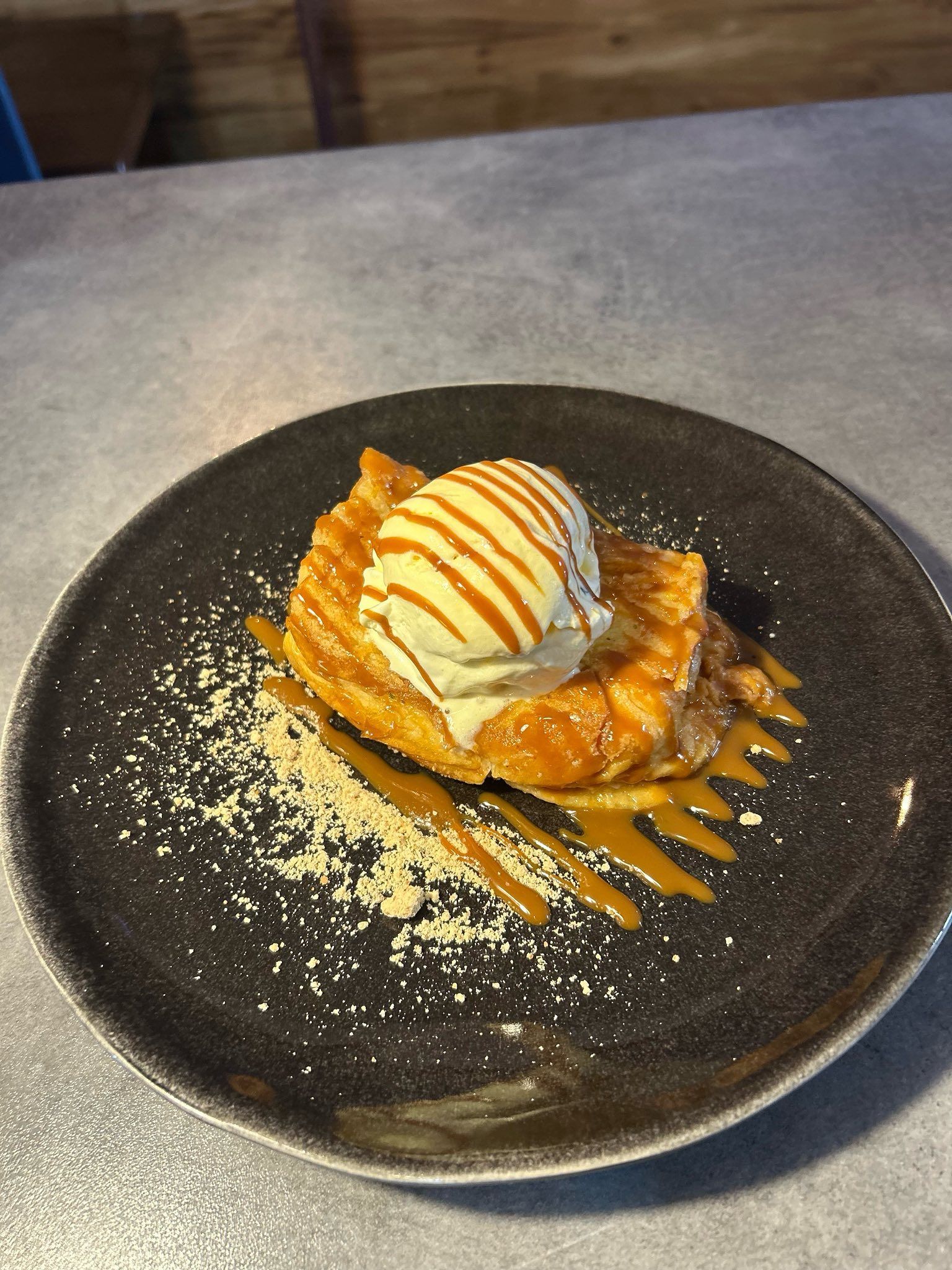 Postre: Salsa de caramelo rociada sobre una masa con helado de vainilla y chispas en un plato oscuro.