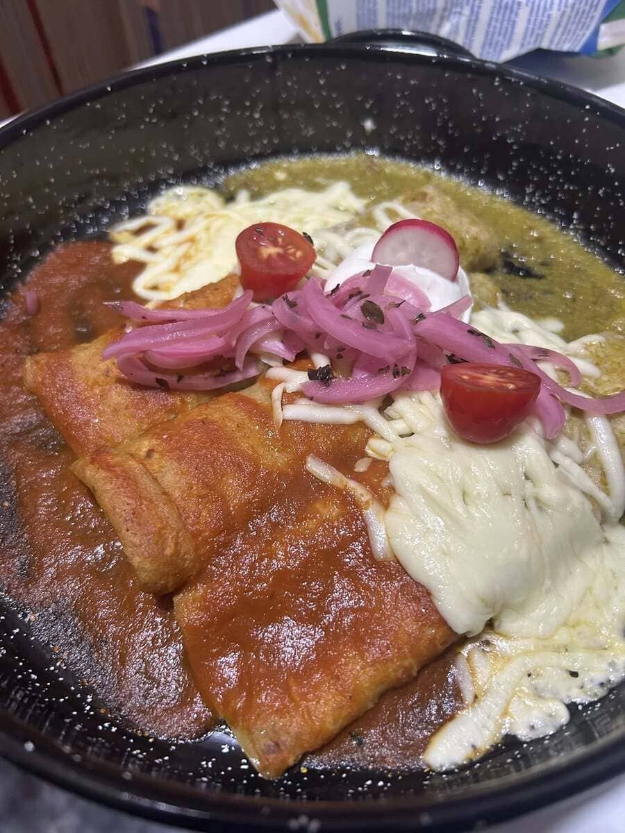Enchiladas en sartén negra, cubiertas con salsa roja y verde, queso, cebollas encurtidas, tomates y crema agria.