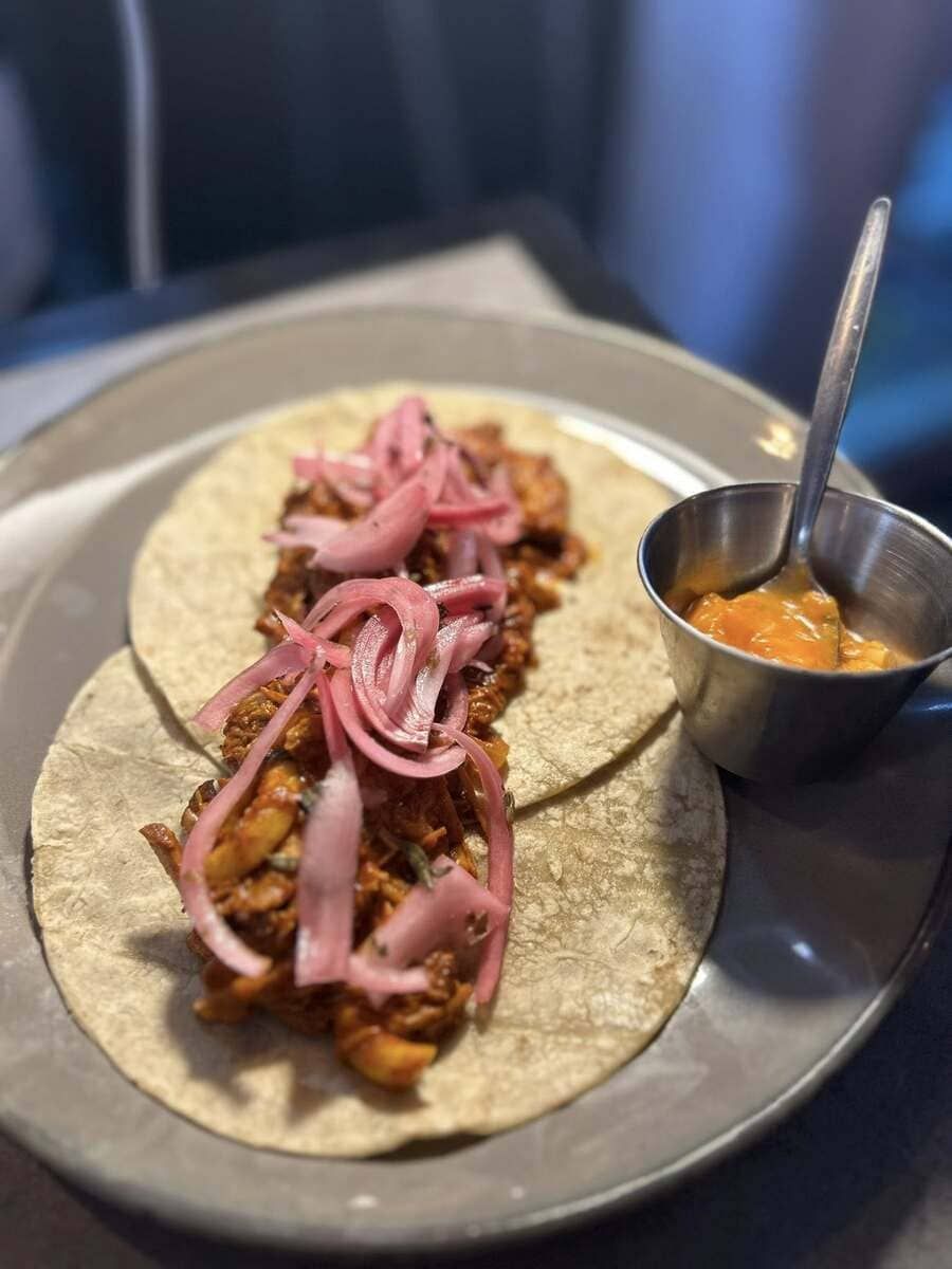 Dos tacos cubiertos con cebollas rojas, junto a un pequeño plato de salsa de naranja en un plato.
